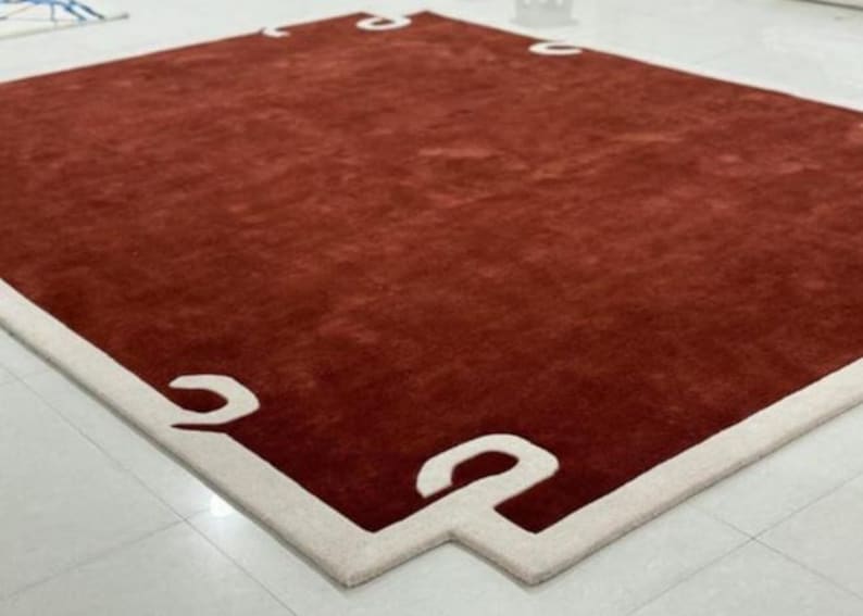 Puede incluir: Una alfombra roja con un borde blanco que presenta un dise&ntilde;o geom&eacute;trico. La alfombra tiene una textura suave y mullida.