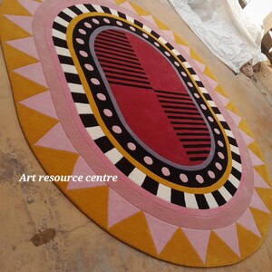May include: A large, oval-shaped rug with a colorful geometric design. The rug features a central oval with black and white stripes, surrounded by a ring of pink and black circles. The outer edge of the rug is a yellow and pink sunburst pattern. The text "Art resource centre" is visible in the lower left corner of the rug.