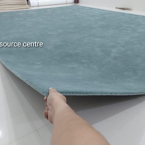 Può includere: Un grande tappeto rettangolare di colore verde acqua tinta unita con una consistenza morbida e soffice. Il tappeto presenta angoli arrotondati e la scritta Art resource centre stampata in bianco sul lato sinistro della superficie.