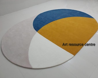 May include: A round rug with a geometric design in shades of white, light pink, blue, and yellow. The rug has a curved shape and features a large yellow section with a blue crescent shape on top.