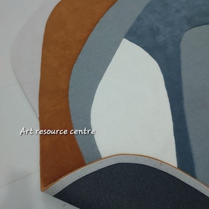 Puede incluir: Primer plano de una alfombra con un dise&ntilde;o geom&eacute;trico en tonos marrones, grises y blancos. La alfombra tiene un respaldo negro y un borde blanco. El texto "Art resource centre" es visible en la esquina inferior izquierda de la imagen.