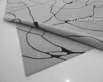 Puede incluir: Una alfombra gris claro con un diseño moderno. La alfombra presenta un patrón de líneas negras irregulares que crean formas abstractas. La alfombra está parcialmente enrollada, revelando la parte inferior. El texto "Art resource centre" es visible en la alfombra.