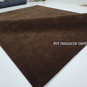 Op de afbeelding: Een groot, vierkant, effen donkerbruin tapijt met een zachte, gelijkmatige textuur. Het tapijt is op één hoek omgevouwen, waardoor de dikte en de pool zichtbaar zijn. De tekst 'Art resource centre' is in het wit op het tapijt gedrukt.