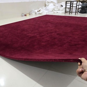 May include: A large, rectangular area rug in a deep, solid burgundy color. The rug features a thick, plush pile texture with a finished, bound edge that is being lifted slightly to display the density and thickness of the material.