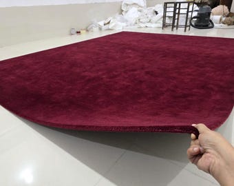 Puede incluir: Una alfombra rectangular grande de color burdeos sólido y profundo. La alfombra presenta una textura de pelo grueso y afelpado con un borde acabado y rematado que se levanta ligeramente para mostrar la densidad y el grosor del material.