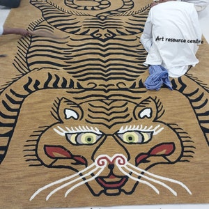 May include: A large, intricately woven rug depicting a tiger with black and white stripes. The rug is a golden brown color and has a detailed face with green eyes. The rug is being laid out on a white tiled floor by several people. The text "Art resource centre" is visible in the image.