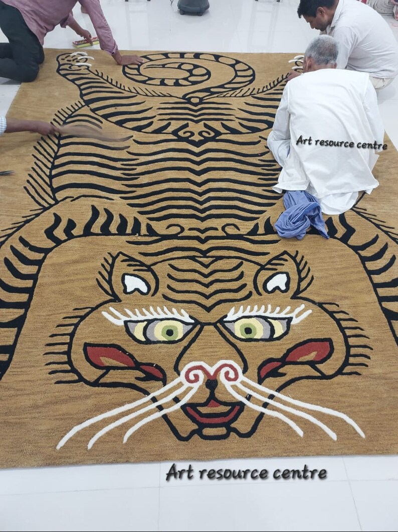 May include: A large, intricately woven rug depicting a tiger with black and white stripes. The rug is a golden brown color and has a detailed face with green eyes. The rug is being laid out on a white tiled floor by several people. The text "Art resource centre" is visible in the image.