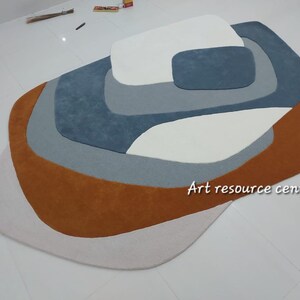 Puede incluir: Una alfombra abstracta multicolor con un dise&ntilde;o en capas. La alfombra presenta tonos de marr&oacute;n, blanco, gris y azul. La alfombra est&aacute; hecha de un material suave y mullido. "Art resource centre" est&aacute; impreso en la alfombra.