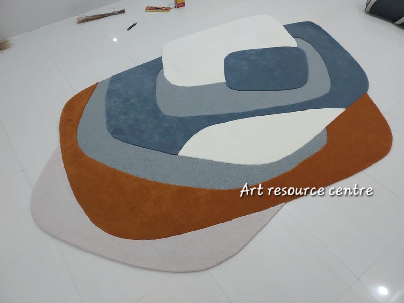 Puede incluir: Una alfombra abstracta multicolor con un dise&ntilde;o en capas. La alfombra presenta tonos de marr&oacute;n, blanco, gris y azul. La alfombra est&aacute; hecha de un material suave y mullido. "Art resource centre" est&aacute; impreso en la alfombra.