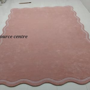 Puede incluir: Una alfombra rosa con un borde blanco festoneado. La alfombra tiene una textura suave y mullida. "Art resource centre" est&aacute; escrito en negro en la alfombra.