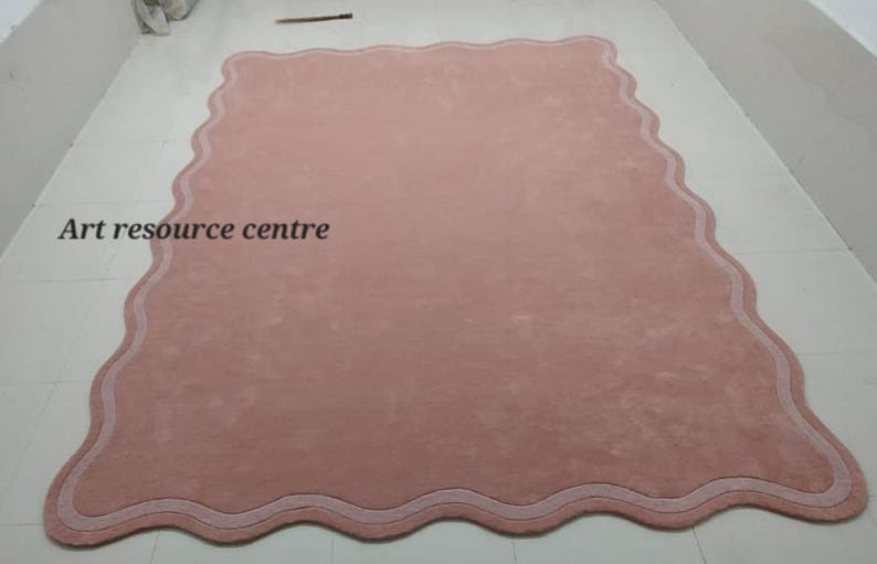 Puede incluir: Una alfombra rosa con un borde blanco festoneado. La alfombra tiene una textura suave y mullida. "Art resource centre" est&aacute; escrito en negro en la alfombra.