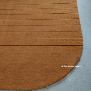 Puede incluir: Una alfombra semicircular de color marr&oacute;n anaranjado con detalles acanalados horizontales. La alfombra est&aacute; hecha de un material suave y afelpado y est&aacute; colocada sobre un suelo de baldosas de color claro. El texto "Art resource centre" es visible en la alfombra.