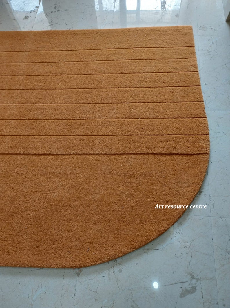 Puede incluir: Una alfombra semicircular de color marr&oacute;n anaranjado con detalles acanalados horizontales. La alfombra est&aacute; hecha de un material suave y afelpado y est&aacute; colocada sobre un suelo de baldosas de color claro. El texto "Art resource centre" es visible en la alfombra.