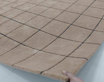 May include: A large, beige rug with a grid pattern of squares and dark blue lines. The rug is being held up at the edge, showing its thickness. The text "Art resource centre" is visible on the rug.