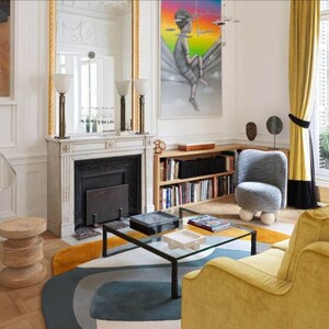Puede incluir: Un interior de sala de estar con un sof&aacute; de terciopelo amarillo, un sill&oacute;n gris, una mesa de caf&eacute; de vidrio y una alfombra con formas abstractas en tonos de azul, gris y blanco. La habitaci&oacute;n cuenta con una chimenea, una estanter&iacute;a y una gran pintura con un fondo de arco&iacute;ris.