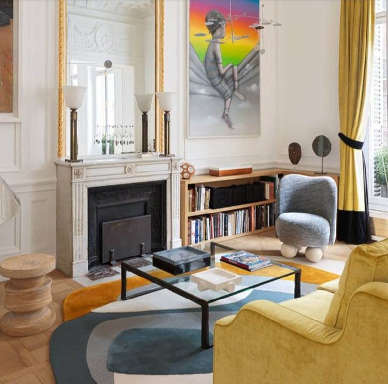 Puede incluir: Un interior de sala de estar con un sof&aacute; de terciopelo amarillo, un sill&oacute;n gris, una mesa de caf&eacute; de vidrio y una alfombra con formas abstractas en tonos de azul, gris y blanco. La habitaci&oacute;n cuenta con una chimenea, una estanter&iacute;a y una gran pintura con un fondo de arco&iacute;ris.