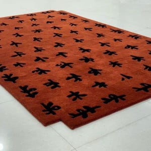 Puede incluir: Primer plano de una alfombra grande y rectangular con un patr&oacute;n repetitivo de hojas estilizadas negras sobre un fondo naranja quemado.