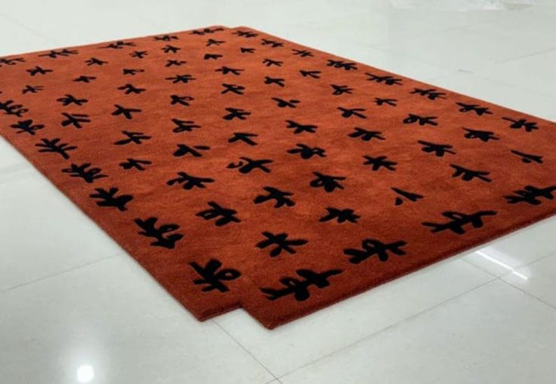 Puede incluir: Primer plano de una alfombra grande y rectangular con un patr&oacute;n repetitivo de hojas estilizadas negras sobre un fondo naranja quemado.