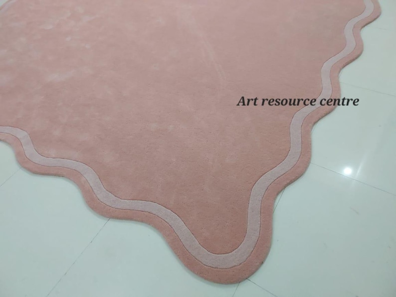 Puede incluir: Una alfombra rosa con un borde festoneado y un borde blanco. La alfombra est&aacute; hecha de un material suave y mullido.