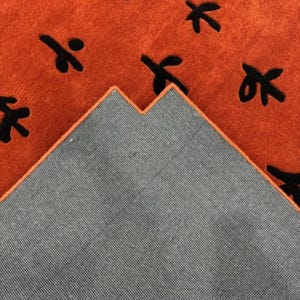 Puede incluir: Primer plano de una alfombra con un respaldo gris oscuro y una parte superior naranja. La parte superior naranja tiene un patr&oacute;n de hojas estilizadas negras.