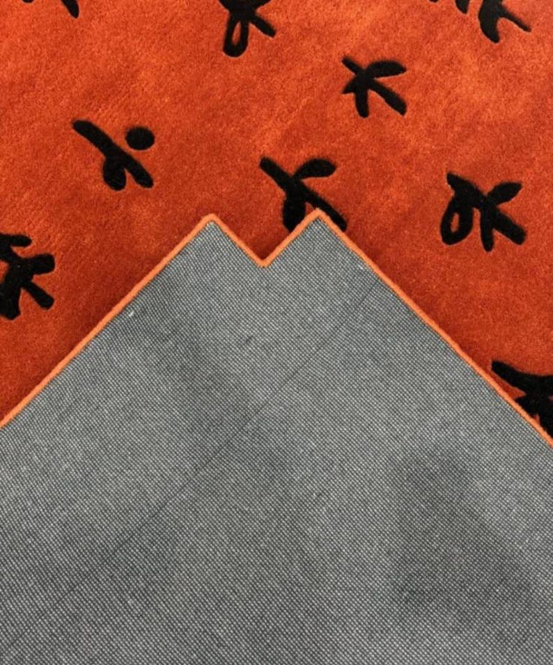 Puede incluir: Primer plano de una alfombra con un respaldo gris oscuro y una parte superior naranja. La parte superior naranja tiene un patr&oacute;n de hojas estilizadas negras.