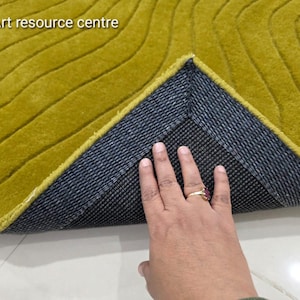 Puede incluir: Primer plano de una alfombra de color amarillo mostaza con un patrón texturizado en forma de onda. La parte inferior de la alfombra es de color gris oscuro con una textura tejida. El texto "Art resource centre" está en la esquina superior izquierda.