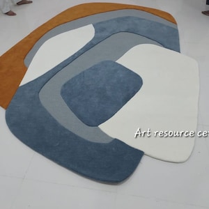 Puede incluir: Alfombra de &aacute;rea abstracta con un dise&ntilde;o moderno que presenta una combinaci&oacute;n de colores naranja, blanco, gris y azul. La alfombra tiene una forma org&aacute;nica &uacute;nica y una textura suave y mullida. "Art resource centre" est&aacute; escrito en texto blanco en la alfombra.