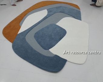 May include: Abstract area rug with a modern design featuring a combination of orange, white, gray, and blue colors. The rug has a unique, organic shape and a soft, plush texture. "Art resource centre" is written in white text on the rug.