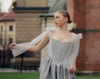 May include: A light gray mini dress featuring a sweetheart neckline, a shimmering silver sequined bodice, and a smooth satin skirt. The dress includes sheer tulle shoulder straps that drape down the arms, paired with a simple white pearl necklace.