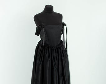 May include: A floor-length black dress featuring a structured bodice with a straight neckline and thin ribbon shoulder ties. The skirt is gathered at the waist, creating a full, voluminous silhouette with a smooth, satin-like finish throughout.
