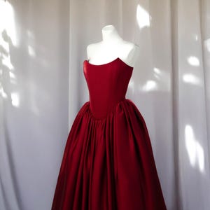 May include: A strapless, floor-length red gown displayed on a white mannequin. The dress features a fitted bodice and a full, flowing skirt. The fabric has a satin sheen, and the dress is set against a white backdrop.