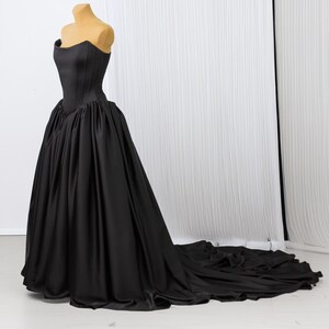 Könnte beinhalten: Ein trägerloses schwarzes Kleid mit eng anliegendem Oberteil und weitem Rock, präsentiert auf einer Schaufensterpuppe. Das Kleid hat eine lange Schleppe und besteht aus einem glatten, satinartigen Stoff. Der Hintergrund ist ein weißer Vorhang.