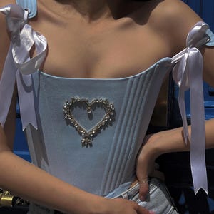 May include: Light blue corset top with a heart-shaped embellishment. The corset features vertical seams, ribbon straps, and white bows at the shoulders. The heart-shaped embellishment is made of silver-colored rhinestones.
