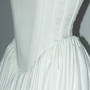 May include: A white corset-style bodice featuring vertical boning channels and a smooth, matte finish. The bodice connects to a voluminous, gathered skirt with deep, vertical pleats that create a full, draped silhouette throughout the garment.