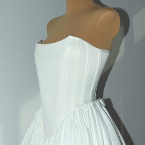 May include: A white strapless corset bodice dress featuring a structured, boned top with vertical seams and a voluminous, gathered skirt. The garment is displayed on a tan mannequin, showcasing a smooth, satin-like fabric finish throughout.