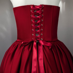 May include: A deep red corset bodice featuring a structured back with silver-toned metal eyelets and a matching satin ribbon lace-up closure. The garment is attached to a full, pleated skirt in the same deep red fabric, tied with a bow.