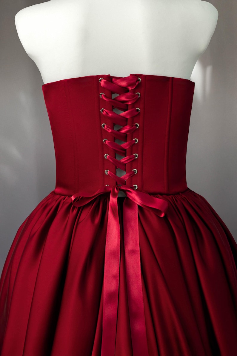 May include: A deep red corset bodice featuring a structured back with silver-toned metal eyelets and a matching satin ribbon lace-up closure. The garment is attached to a full, pleated skirt in the same deep red fabric, tied with a bow.