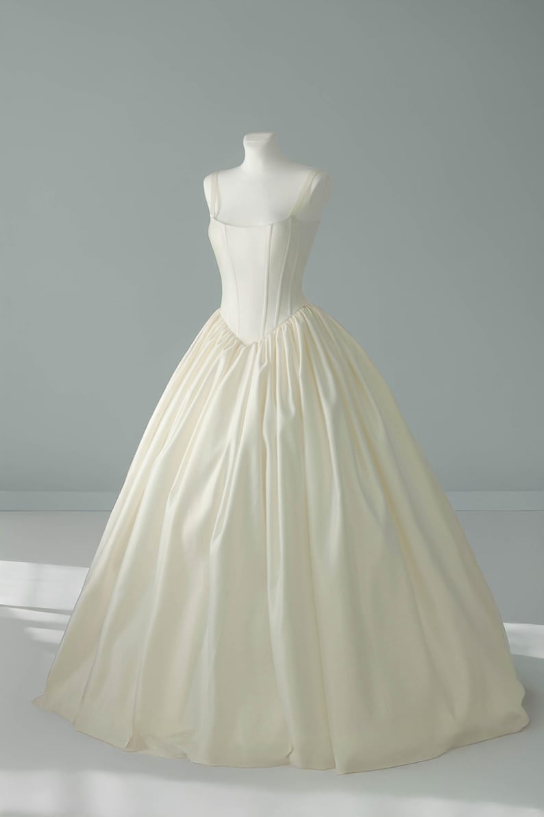 May include: A floor-length cream-colored gown featuring a structured, corset-style bodice with vertical seams and a square neckline. The dress has wide shoulder straps and a voluminous, gathered skirt that flares out from the waistline.