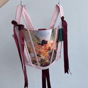 Puede incluir: Un cors&eacute; rosa con un dise&ntilde;o estampado de pintura cl&aacute;sica. El cors&eacute; tiene correas rosas y cintas burdeos atadas con lazos. La prenda est&aacute; colgada de una percha plateada.