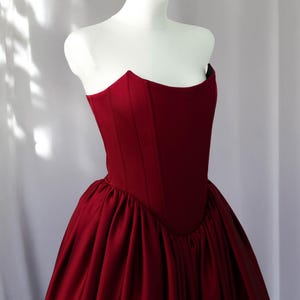 May include: A deep red strapless corset bodice dress featuring a structured, fitted top with vertical seams and a voluminous, gathered skirt. The garment is made of a smooth, solid-coloured fabric and is displayed on a white mannequin torso.