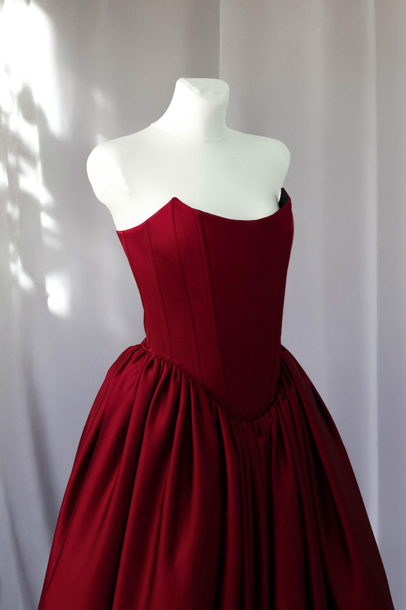 May include: A deep red strapless corset bodice dress featuring a structured, fitted top with vertical seams and a voluminous, gathered skirt. The garment is made of a smooth, solid-coloured fabric and is displayed on a white mannequin torso.