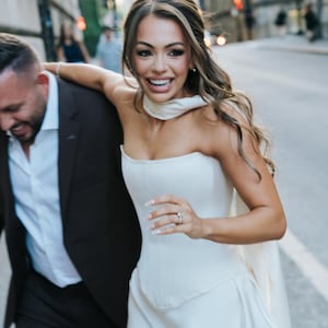 May include: A woman in a white strapless dress with a matching scarf and a ring on her finger. She is smiling and running down a city street with a man in a dark suit. The background features buildings and a blurred street scene.