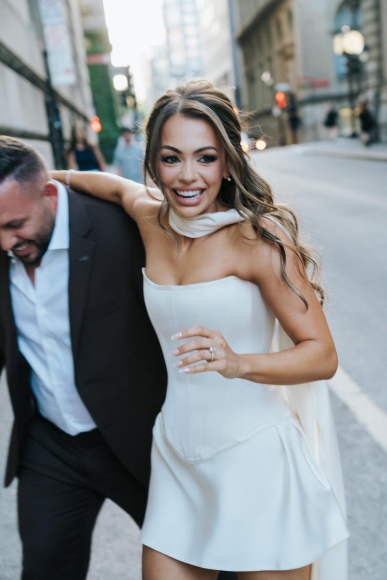 May include: A woman in a white strapless dress with a matching scarf and a ring on her finger. She is smiling and running down a city street with a man in a dark suit. The background features buildings and a blurred street scene.