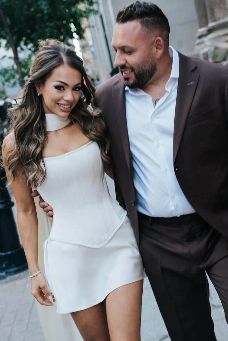 May include: A woman in a white corset-style mini dress with a matching choker. She is smiling and walking next to a man wearing a brown suit jacket over a white button-down shirt. The background features a city street with buildings.