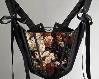 May include: A black corset top featuring a central panel with a dark, multi-coloured floral print of roses and other blossoms. The garment includes black satin shoulder straps that tie into bows and a black lace-up closure at the centre back.