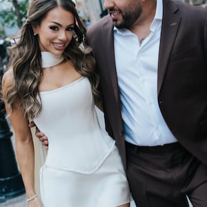 May include: A white corset-style mini dress featuring a structured bodice with vertical seams, a straight neckline, and a matching choker collar. The garment is paired with a dark brown suit jacket and a crisp white button-down shirt.