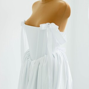 May include: A white, strapless dress with a corset-style bodice and a flowing skirt. The dress features delicate white bows on the shoulders and is displayed on a mannequin. The fabric appears to be lightweight and airy, suitable for formal occasions.