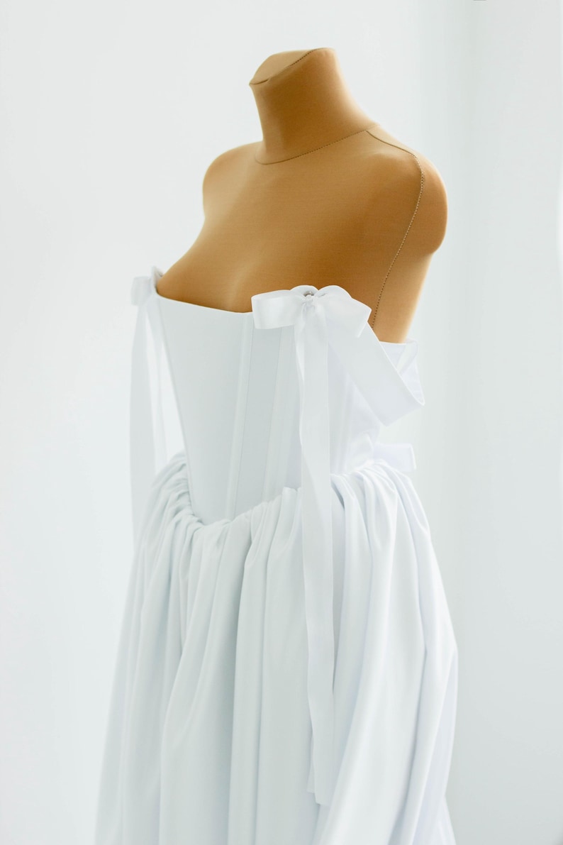 May include: A white, strapless dress with a corset-style bodice and a flowing skirt. The dress features delicate white bows on the shoulders and is displayed on a mannequin. The fabric appears to be lightweight and airy, suitable for formal occasions.
