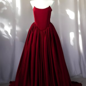 May include: A floor-length, deep red gown featuring a structured, corset-style bodice with a straight neckline and a voluminous, pleated skirt that cascades to the floor. The garment is displayed on a white mannequin, showcasing its solid colour.