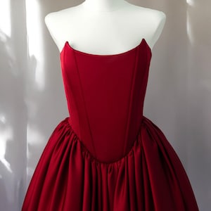 May include: A deep red strapless gown featuring a structured corset-style bodice with vertical seams and a voluminous, gathered skirt. The garment is displayed on a white mannequin, showcasing a smooth, matte finish on the bodice and fabric.